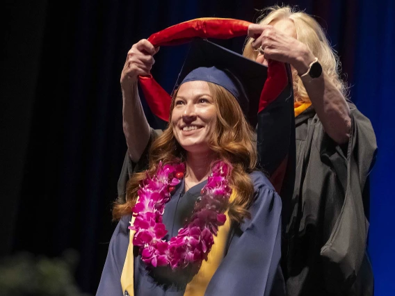 A young woman wearing graduation regalia smiles as a professor places a traditional hood over her shoulders from behind. 