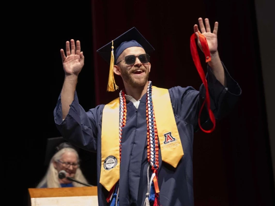 A young man wearing a graduation cap and gown and sunglasses raises his hands in the air as he walks across a stage. 