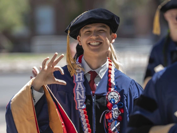 A young man in graduation regalia smiles and gives the hand sign for the University of Arizona Wildcats as he walks outside. 