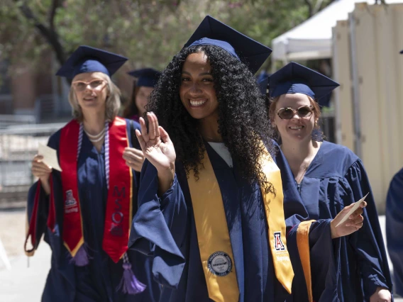 Three young women dressed in graduation caps and gowns smile as they walk in a line outside. 