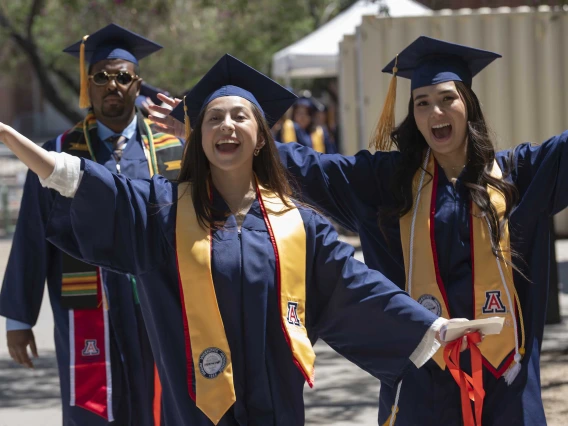 Two young women wearing graduation caps and gowns raise their hands in celebration as they walk outside with other graduates behind them. 