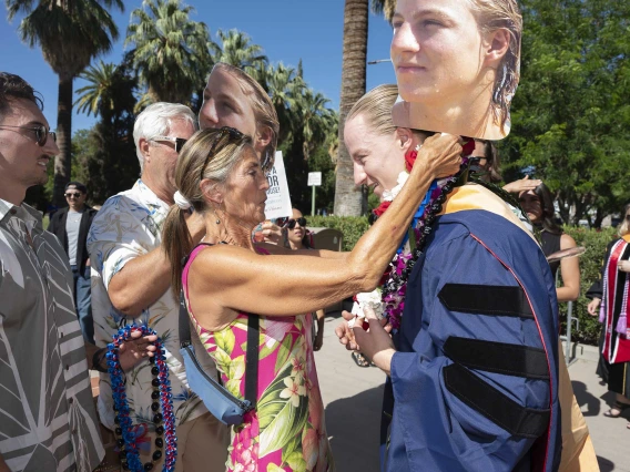 A mother places a flower lei around her son’s neck while holding a large photo of him in her hand. Her son is wearing a graduation gown. 
