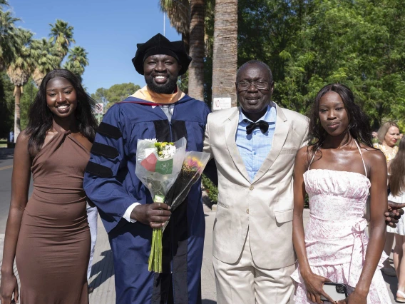 A young man in graduation regalia holds a bouquet of flowers and smiles as he stands with his two sisters and father outside. 