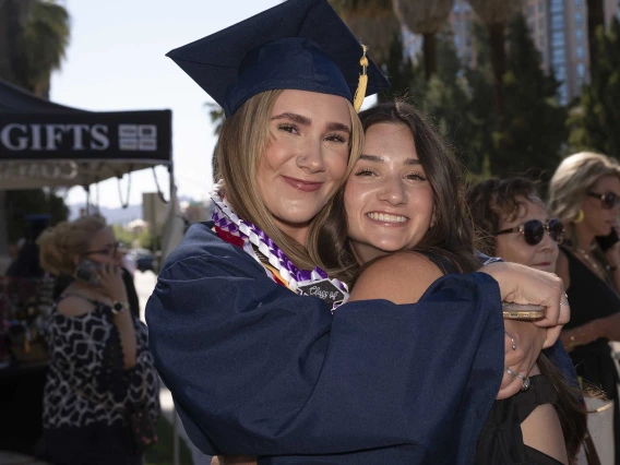 A young woman wearing a graduation cap and gown smiles as she hugs another smiling young woman. 