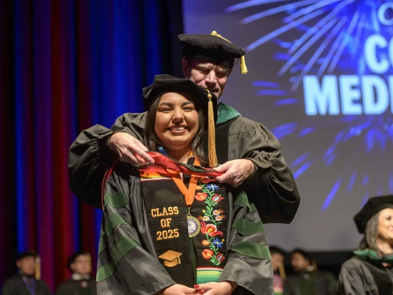 A medical student in a graduation cap and gown has a ceremonial hood placed over her shoulders by a professor in similar attire. 