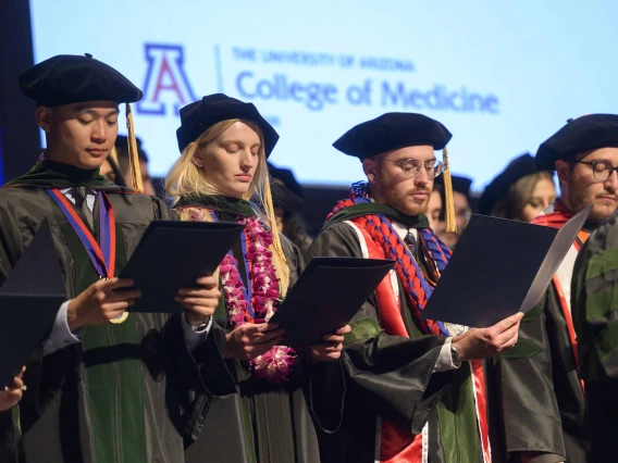 Several medical students in graduation caps and gowns read an oath from folders they are holding on a stage. 