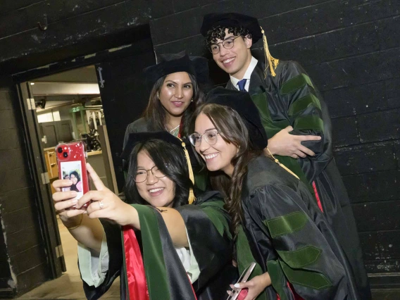 Four medical students in graduation caps and gowns bunch together to take a selfie. 