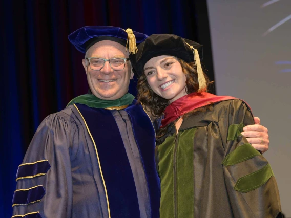 A father and daughter wearing graduation regalia smile and side hug during a graduation ceremony. 