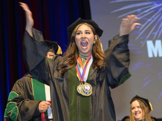 A young woman dressed in graduation regalia raises her arms in celebration with her mouth open, cheering. 