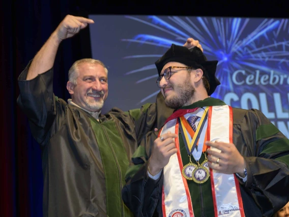 A father points in celebration at his son, both wearing graduation regalia, during a convocation ceremony. 