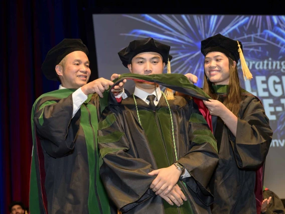 A medical student in a graduation cap and gown stands onstage as his father and sister, also in graduation regalia, place a ceremonial hood over his shoulders. 