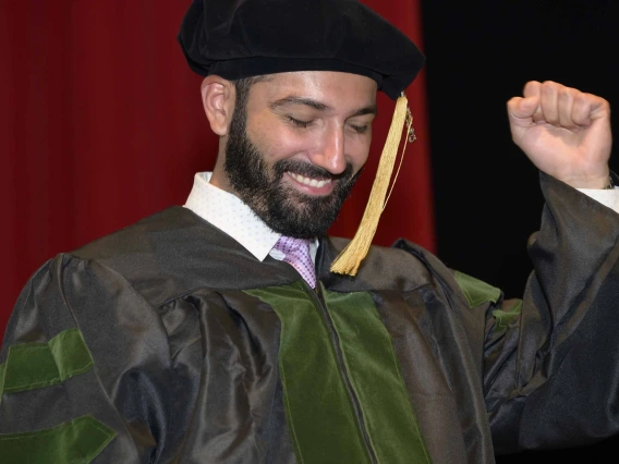 A medical student wearing graduation regalia pumps his fist and smiles onstage during a graduation ceremony. 