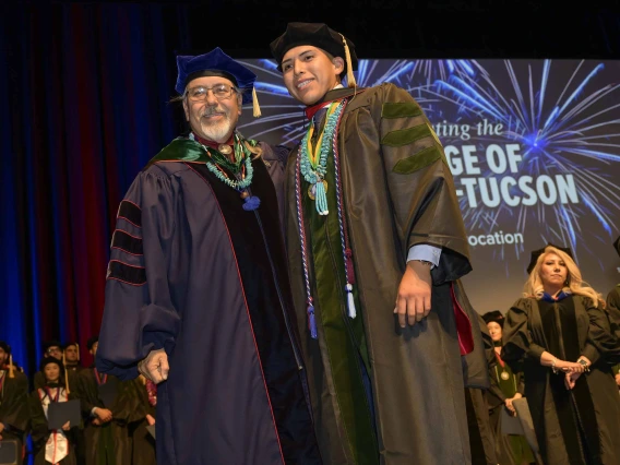 A professor and medical student in graduation regalia stand together onstage during a graduation ceremony. 
