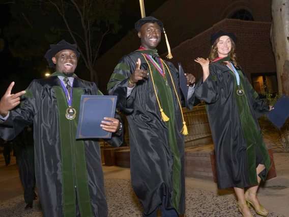 Three medical students in graduation regalia wave in celebration while walking outside at night. 