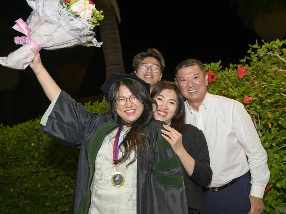 A medical student in a graduation robe holds flowers up in celebration as her smiling family stands behind her. 