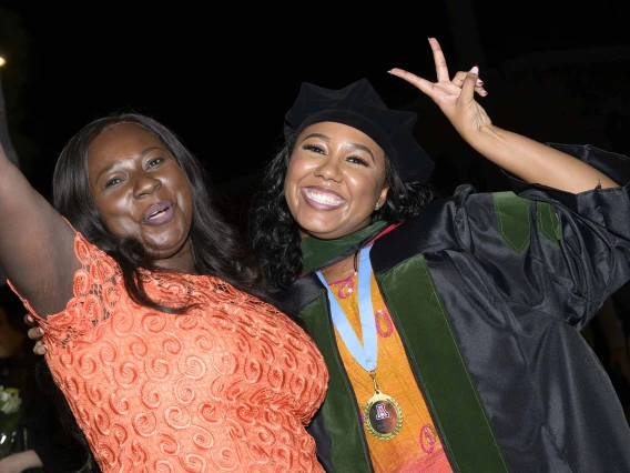 A medical school graduate dressed in a cap and gown, hugs her mother as they raise their hands in the air in celebration. 