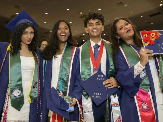 Four college students dressed in caps and gowns smile and cheer at a graduation ceremony. 