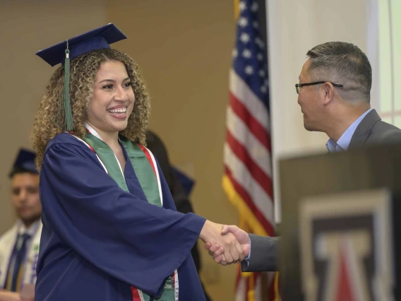 A young woman in a graduation cap and gown smiles as she shakes hands with a man during a graduation ceremony. 