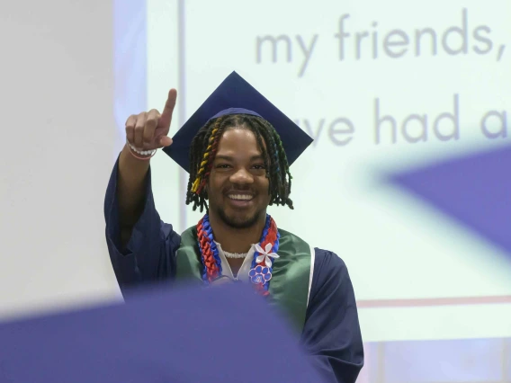 A student wearing graduation regalia stands onstage smiling and holding up a finger in celebration. 