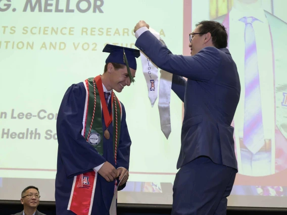 A young man wearing a graduation cap and gown is presented with a white sash onstage by a professor during a graduation ceremony. 