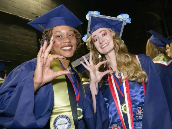 Two young women in graduation caps and gowns smile while giving the hand sign for the Arizona Wildcats. 