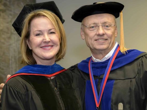 Two professors in graduation regalia stand together, smiling. 