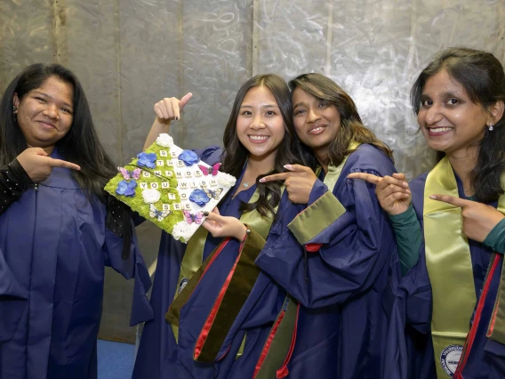 Four young women dressed in graduation regalia smile as they hold up a decorated graduation cap. 
