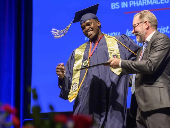 A young man dressed in a graduation cap and gown smiles as he is handed his diploma by an older man in a suit. 