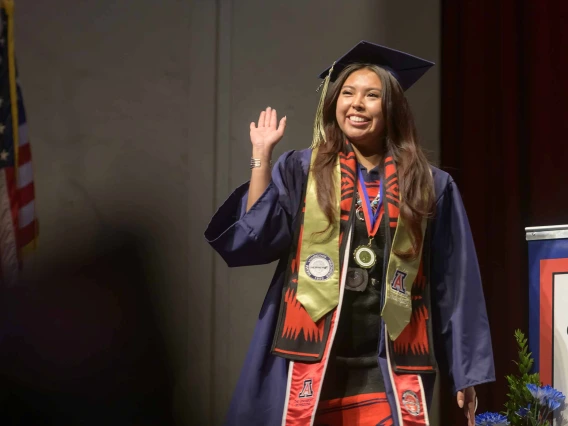 A young woman wearing graduation regalia smiles and waves as she walks onto a stage. 