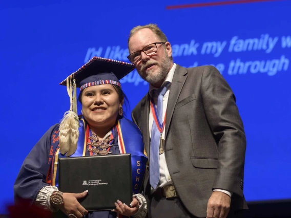 A young woman dressed in a graduation cap and gown holds her diploma and smiles as a professor in a suit stands next to her. 