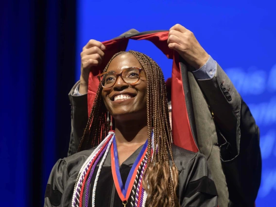 A young woman dressed in a graduation gown smiles as a professor places a ceremonial hood over her shoulders.