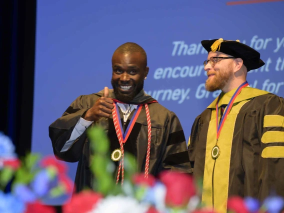 A young man in graduation regalia smiles and gives a thumbs-up while standing next to a professor who is wearing a cap and gown. 