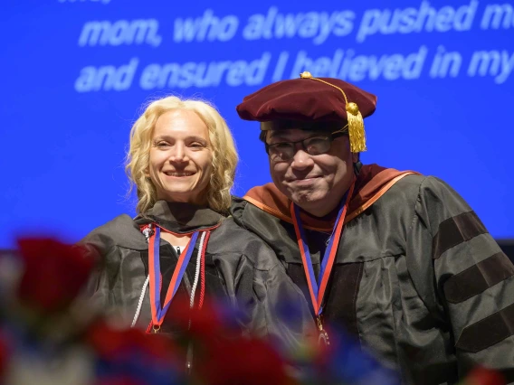 A young woman dressed in a graduation gown smiles as a professor, also dressed in graduation regalia, smiles behind her. 