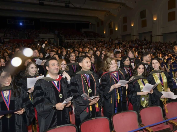 More than 225 students dressed in graduation regalia stand in an auditorium at the conclusion of their convocation ceremony. 