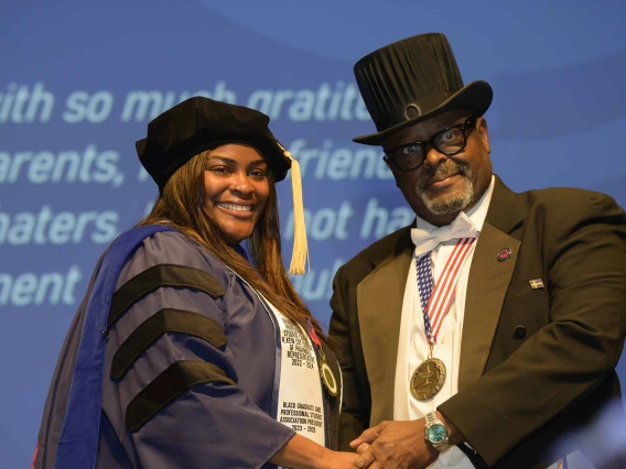 A student in a cap and gown shakes hands with a professor in a suit and top hat during a graduation ceremony. 