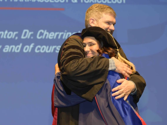 A professor hugs a woman onstage during a graduation ceremony. Both are dressed in graduation regalia. 