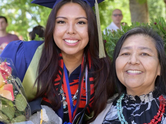 A young woman in a graduation cap and gown holds flowers as she stands next to her aunt in an outdoor setting. 