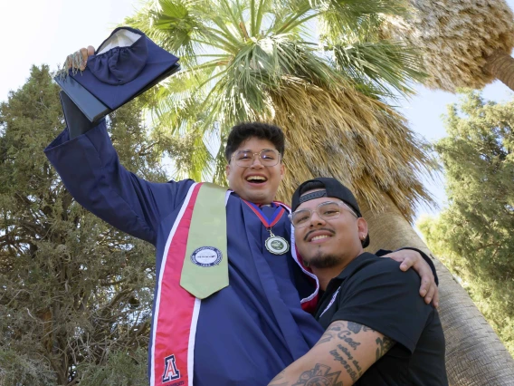 A young man in graduation regalia is hoisted into the air by another young man in celebration. Both are smiling outside under some trees.