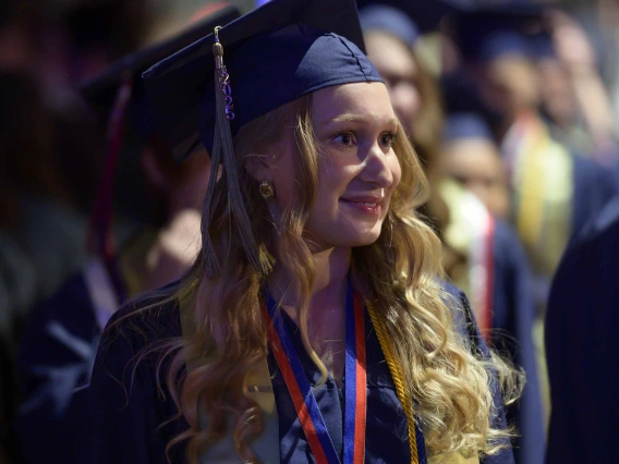 A young woman in a graduation cap and gown smiles as she looks off into the distance with other graduates behind her. 