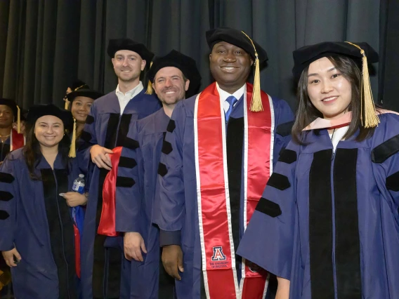 About a dozen doctoral students wearing graduation regalia smile as they stand in line in front of dark stage curtains. 
