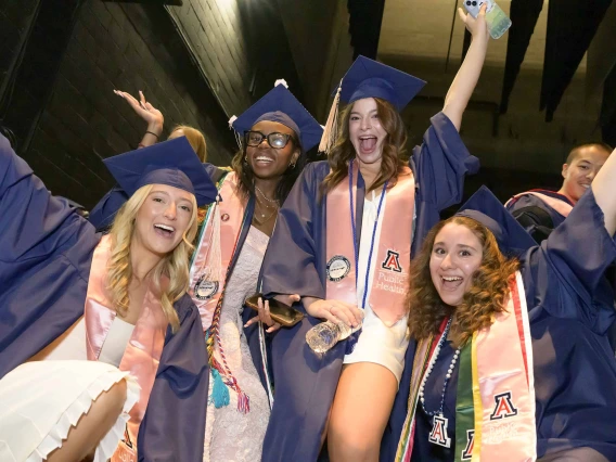 Four young women in graduation caps and gowns raise their hands and cheer in celebration. 