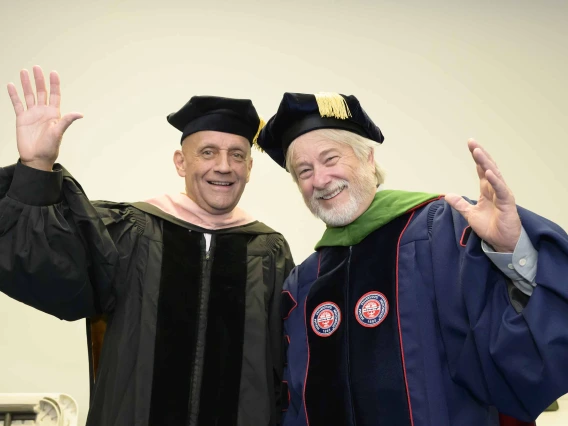 Two professors in graduation regalia smile and wave. 