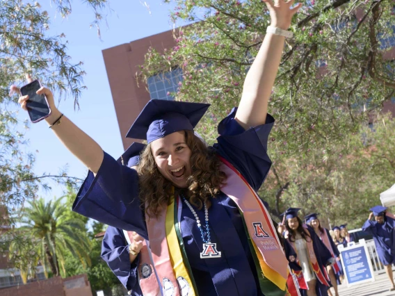 A young woman in a graduation cap and gown raises her arms in celebration as she leads a line of other graduates outside. 