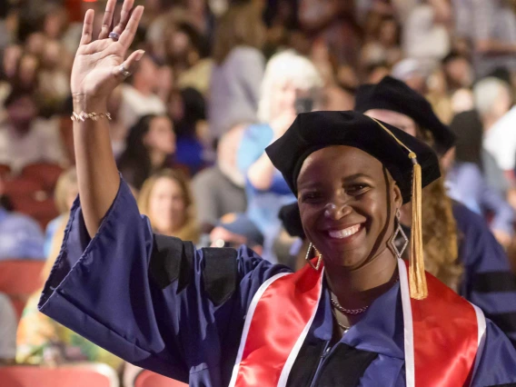 A woman wearing graduation regalia smiles and waves as she walks down an aisle of an auditorium filled with people. 