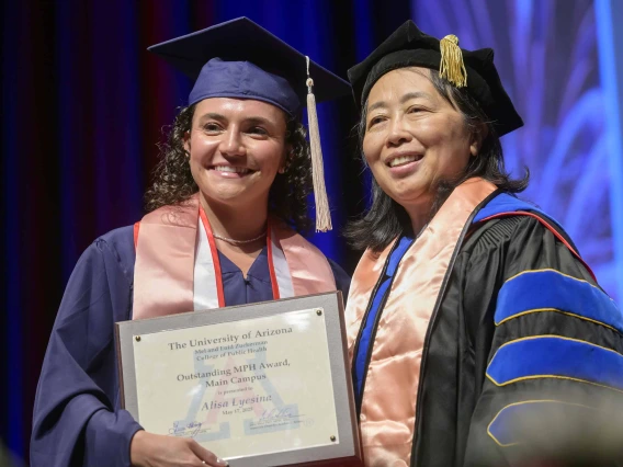 A young woman holding a certificate stands next to a professor onstage during a graduation ceremony. Both are wearing graduation regalia.