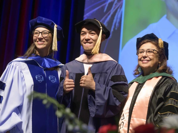A young man gives two thumbs up as he stands between two professors. All are wearing graduation regalia on a stage and there is a projection of the man on a screen behind them. 