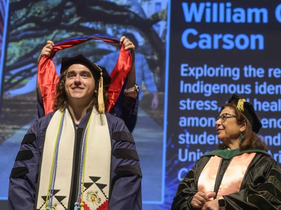 A young man wearing a graduation cap and gown smiles as a professor places a traditional graduation hood over his shoulders. Another professor looks on from the side. 