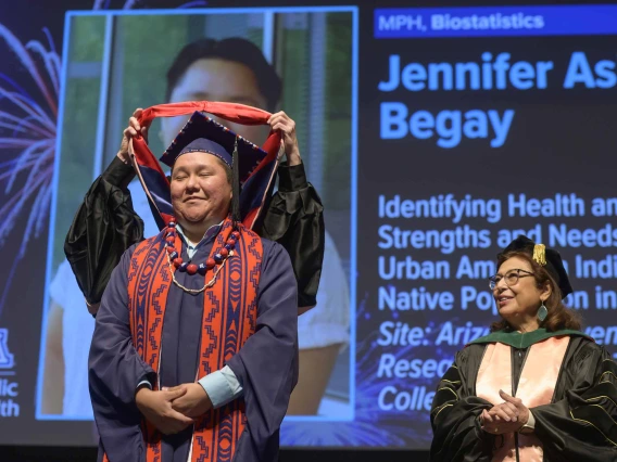 A young woman dressed in a graduation cap and gown closes her eyes and smiles as a professor places a graduation hood over her shoulders while another professor looks on.