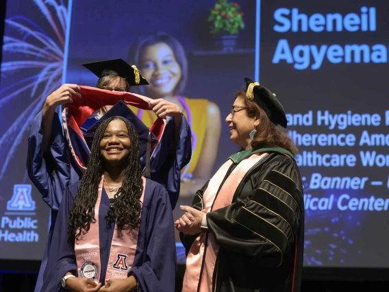 A young woman smiles as a professor places a ceremonial hood over her shoulders during a graduation ceremony while another professor looks on. All are wearing graduation caps and gowns. 
