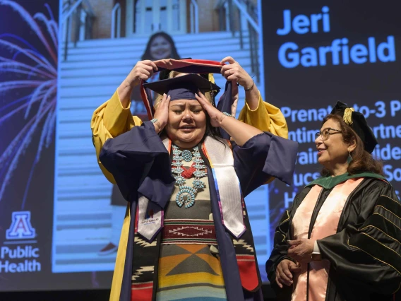 A young woman dressed in a graduation cap and gown and wearing a turquoise necklace and traditional Native American dress holds back tears as a professor places a graduation hood over her shoulders while another professor looks on. 